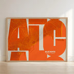 Framed Alicante in Inseparable design, featuring the city's abbreviation in expressive typography over a detailed urban map designed in orange and an off-white background with no border edge-to-edge print leaning against a wall