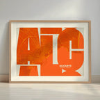 Framed Alicante in Inseparable design, featuring the city's abbreviation in expressive typography over a detailed urban map designed in orange and an off-white background with a white margin border in a warm lit minimalist setting