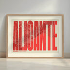 Framed Alicante in Extra Compressed design, where the letters are tightly squeezed to fit the city's map shape designed in red and an off-white background with a white margin border in a warm lit minimalist setting