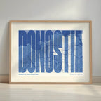 Cuadro de Donostia en el diseño la Extra Comprimida, donde las letras se ajustan al ancho del mapa de la ciudad diseñado en azul marino y blanco con margen en un entorno minimalista con iluminación cálida