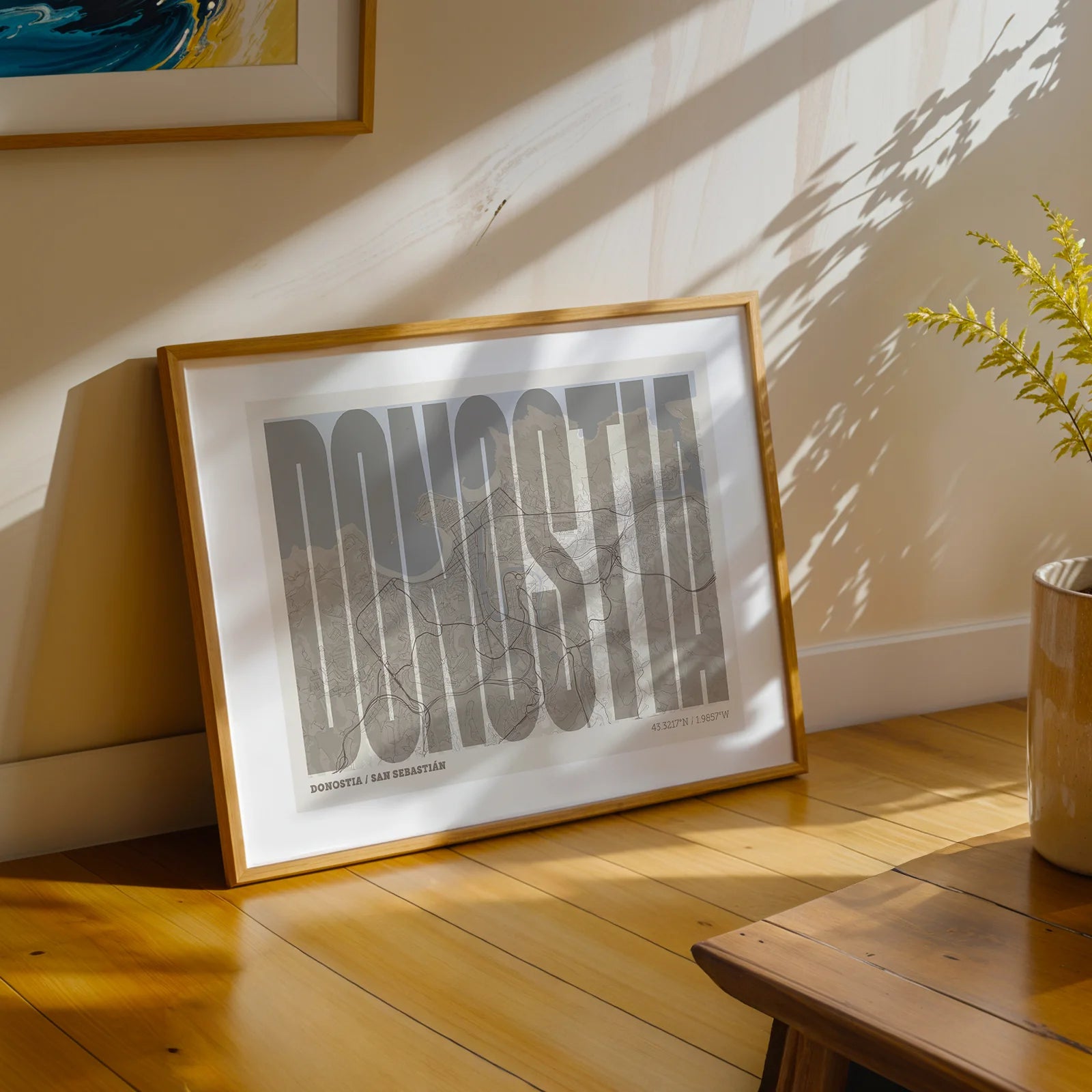 Diseño de interiores moderno con la lámina de Donostia en el diseño la Extra Comprimida, donde las letras se ajustan al ancho del mapa de la ciudad diseñado en marrón claro y fondo blanco roto, expuesto en la pared de un salón
