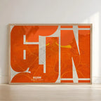 Framed Gijon in Inseparable design, featuring the city's abbreviation in expressive typography over a detailed urban map designed in orange and an off-white background with no border edge-to-edge print leaning against a wall