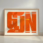 Framed Gijon in Inseparable design, featuring the city's abbreviation in expressive typography over a detailed urban map designed in orange and an off-white background with a white margin border in a warm lit minimalist setting