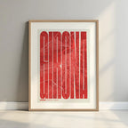 Framed Girona in Extra Compressed design, where the letters are tightly squeezed to fit the city's map shape designed in red and an off-white background with a white margin border in a warm lit minimalist setting