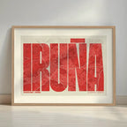 Framed Iruña in Extra Compressed design, where the letters are tightly squeezed to fit the city's map shape designed in red and an off-white background with a white margin border in a warm lit minimalist setting