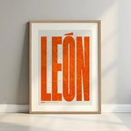 Framed Leon in Extra Compressed design, where the letters are tightly squeezed to fit the city's map shape designed in orange and an off-white background with a white margin border in a warm lit minimalist setting