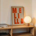 Diseño de interiores moderno con la lámina de Melilla en el diseño la Revoltosa, donde las letras del nombre aparecen aleatoriamente en diferentes líneas para un diseño urbano realmente original diseñado en naranja y fondo blanco roto, expuesto en la pared de un salón