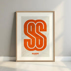 Framed San Sebastian de los Reyes in Super Bold design, a typographic poster where the city map is overlaid with extra-thick lettering for a modern, high-impact look designed in orange and an off-white background with a white margin border in a warm lit minimalist setting