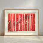Framed San Sebastian in Extra Compressed design, where the letters are tightly squeezed to fit the city's map shape designed in red and an off-white background with a white margin border in a warm lit minimalist setting
