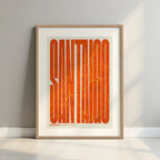 Framed Santiago de Compostela in Extra Compressed design, where the letters are tightly squeezed to fit the city's map shape designed in orange and an off-white background with a white margin border in a warm lit minimalist setting