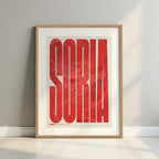 Framed Soria in Extra Compressed design, where the letters are tightly squeezed to fit the city's map shape designed in red and an off-white background with a white margin border in a warm lit minimalist setting