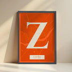 Framed Zamora in Classic design, inspired by chiseled typography with an elegant font that creates a beautiful visual contrast designed in orange and an off-white background with no border edge-to-edge print leaning against a wall